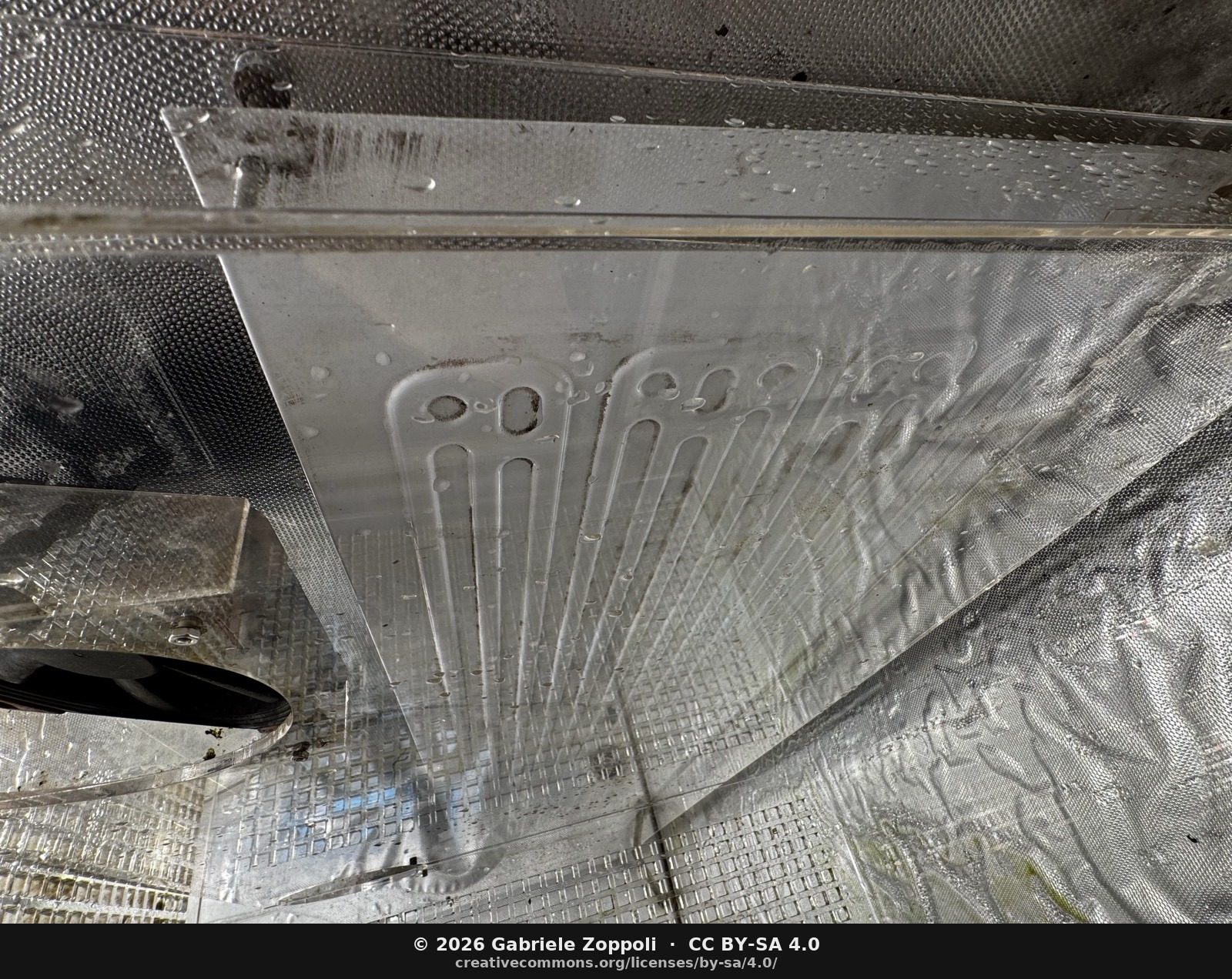 The canalina — the aluminum fold across the bottom of the frame. Suspended on fishing line at 5° toward the drainage pipe; condensate runs off the back of the evaporator (the serpentine refrigerant-tube pattern pressed into the aluminum cooling plate, visible above) onto it, then by gravity through a PVC pipe to the tank under the cabinet.